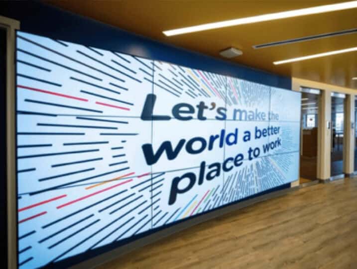 Floor-to-ceiling digital display in an office space displaying the quote “Let’s make the world a better place to work” in large black font with colorful lines around it for emphasis.