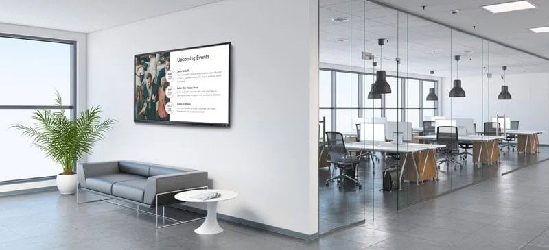 Open office space with a single white wall dividing a seating area from a working space. A gray couch and large plant sit below a digital sign in the seating area. A row of desks and hanging lights occupy the working space.