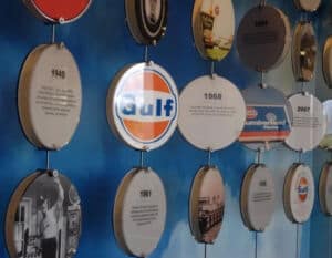 The image features a wall display of various circular plaques, each representing significant milestones or advertisements in the history of the Gulf Oil company. The plaques are a mix of years, images, and logos, with each plaque containing a brief description or image relevant to its era. The centerpiece is the iconic Gulf logo from 1968, displayed prominently among other historical markers. This exhibit serves as a timeline or historical narrative, showcasing the brand's evolution and key moments in its history, likely located in a corporate office or a museum dedicated to the petroleum industry. The design is both informative and visually engaging, providing a snapshot of the company's legacy through decades.